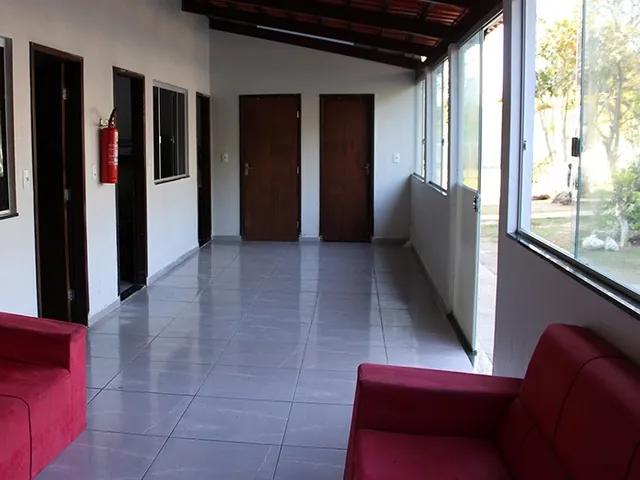 Corredor coberto com piso cerâmico claro, janelas de vidro à direita, portas de madeira à esquerda e ao fundo e dois sofás vermelhos