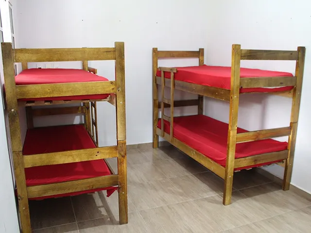 Quarto com paredes brancas e dois beliches de madeira com colchões vermelhos